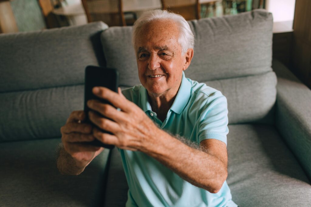 pexels-photo-27086776-27086776 Senior man smiling while using a smartphone on a sofa indoors, expressing joy and engagement.